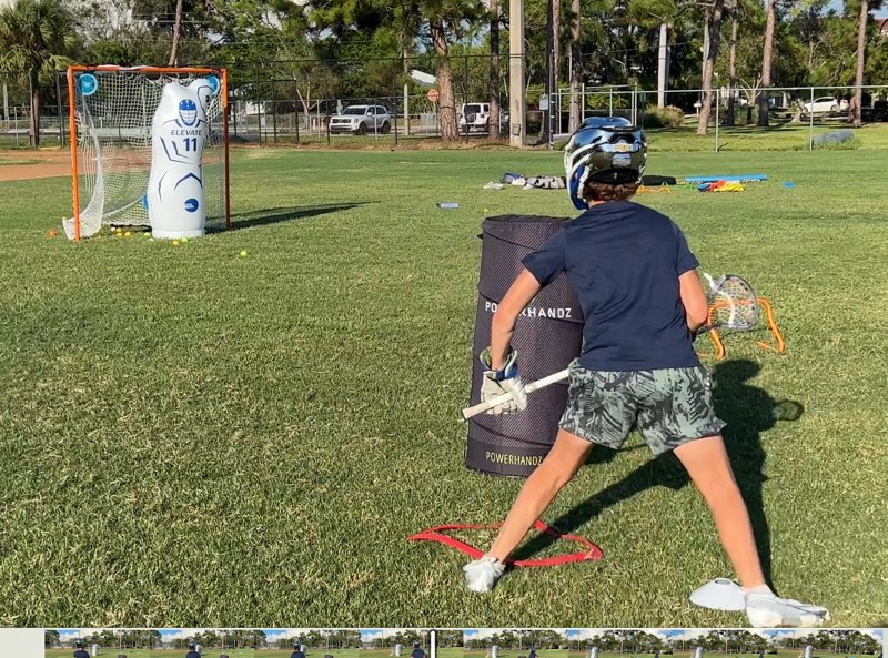Athlete in a helmet practicing a lacrosse shot on a field near a goal.